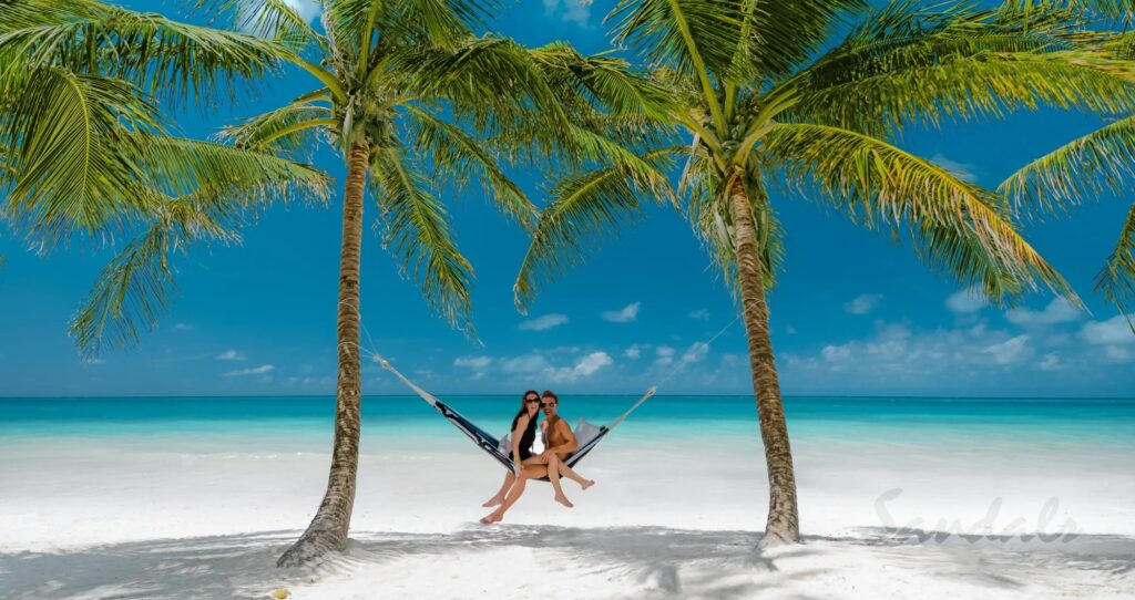 Smiling couple relaxing on a hammock between palm trees, enjoying the picturesque white sand coast and turquoise waters of a luxury Barbados holiday setting_